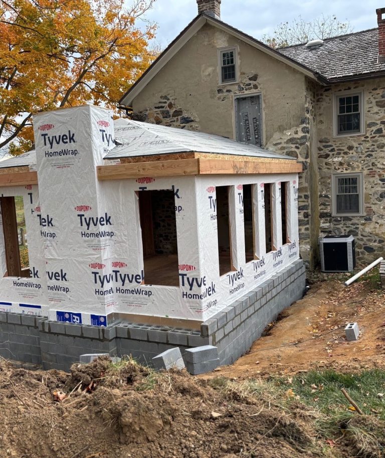 Framed sunroom addition wrapped in weather barrier beside the stone farmhouse