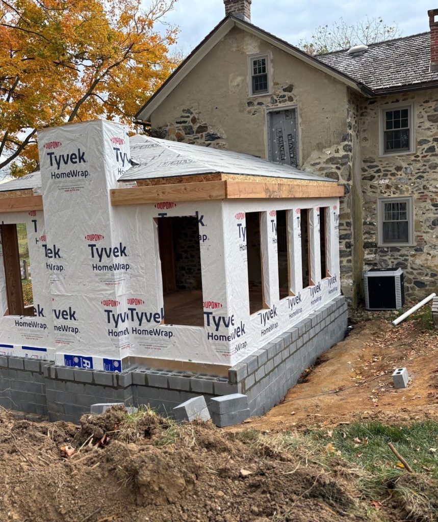 Framed sunroom addition wrapped in weather barrier beside the stone farmhouse